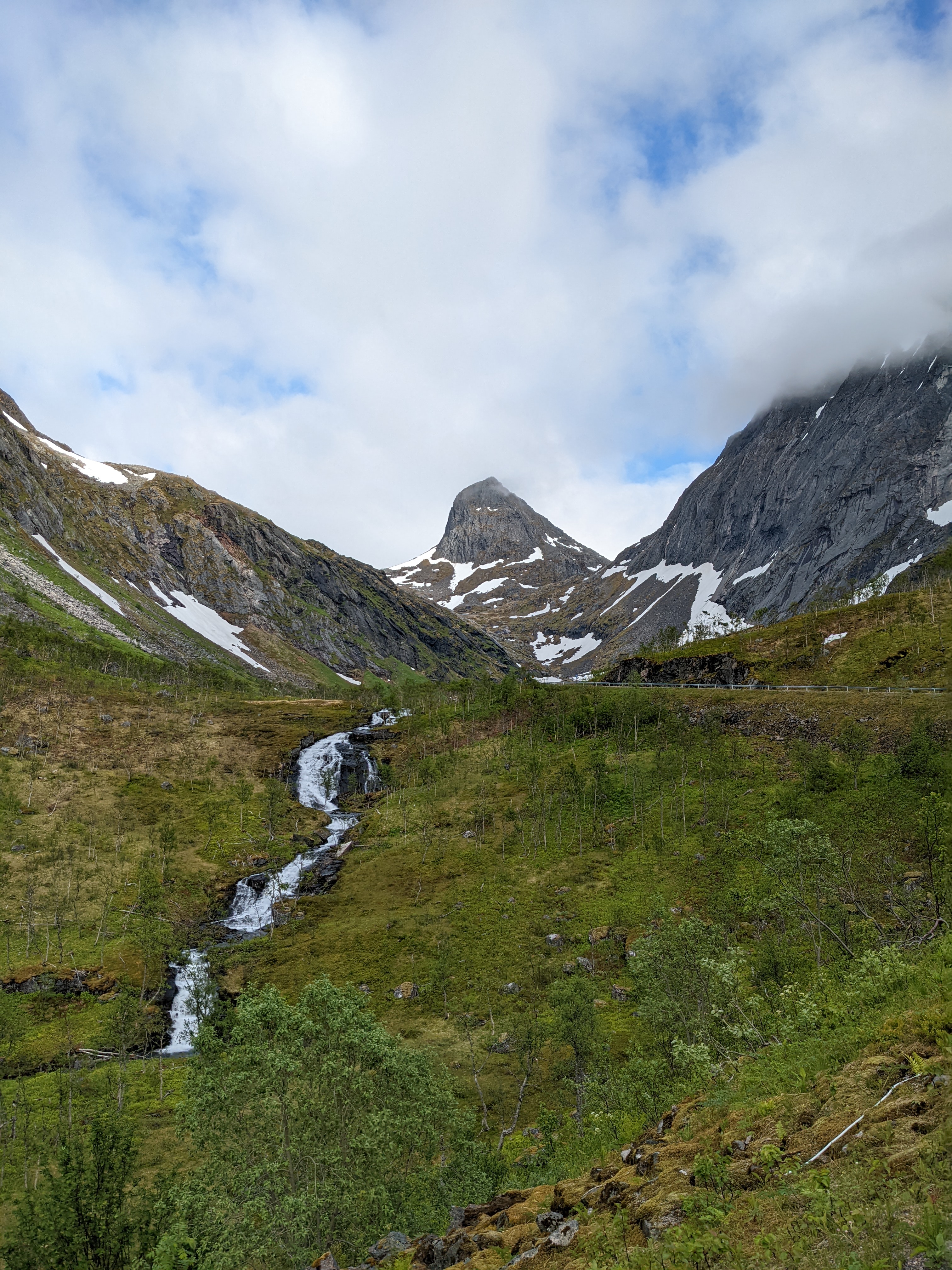 a steep peak above a waterfall and sparse birch trees. the road runs across and up on the right