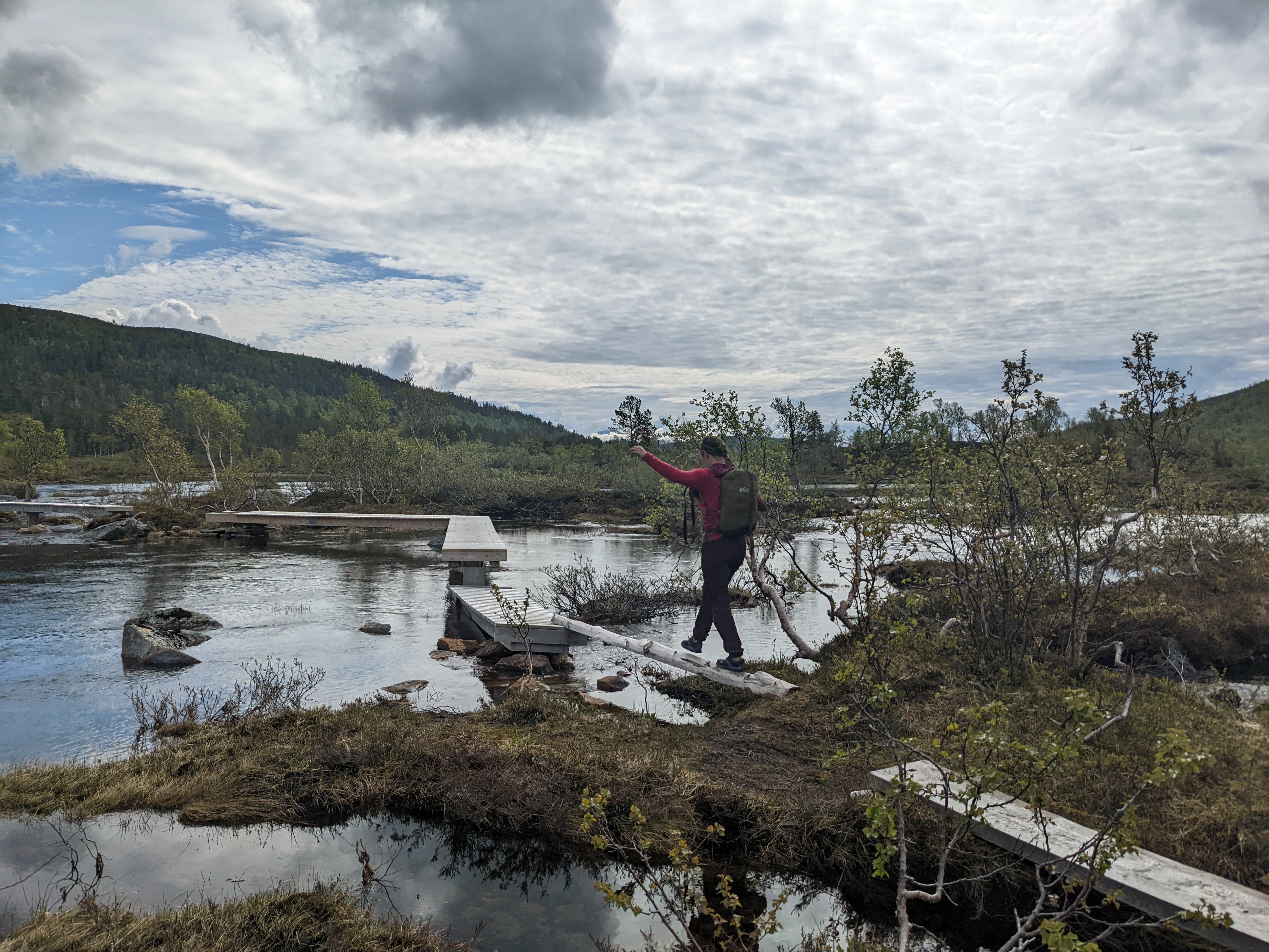 H walking across a log bridge on a boardwalk over the lake outlet, with arms out to the sides for balance