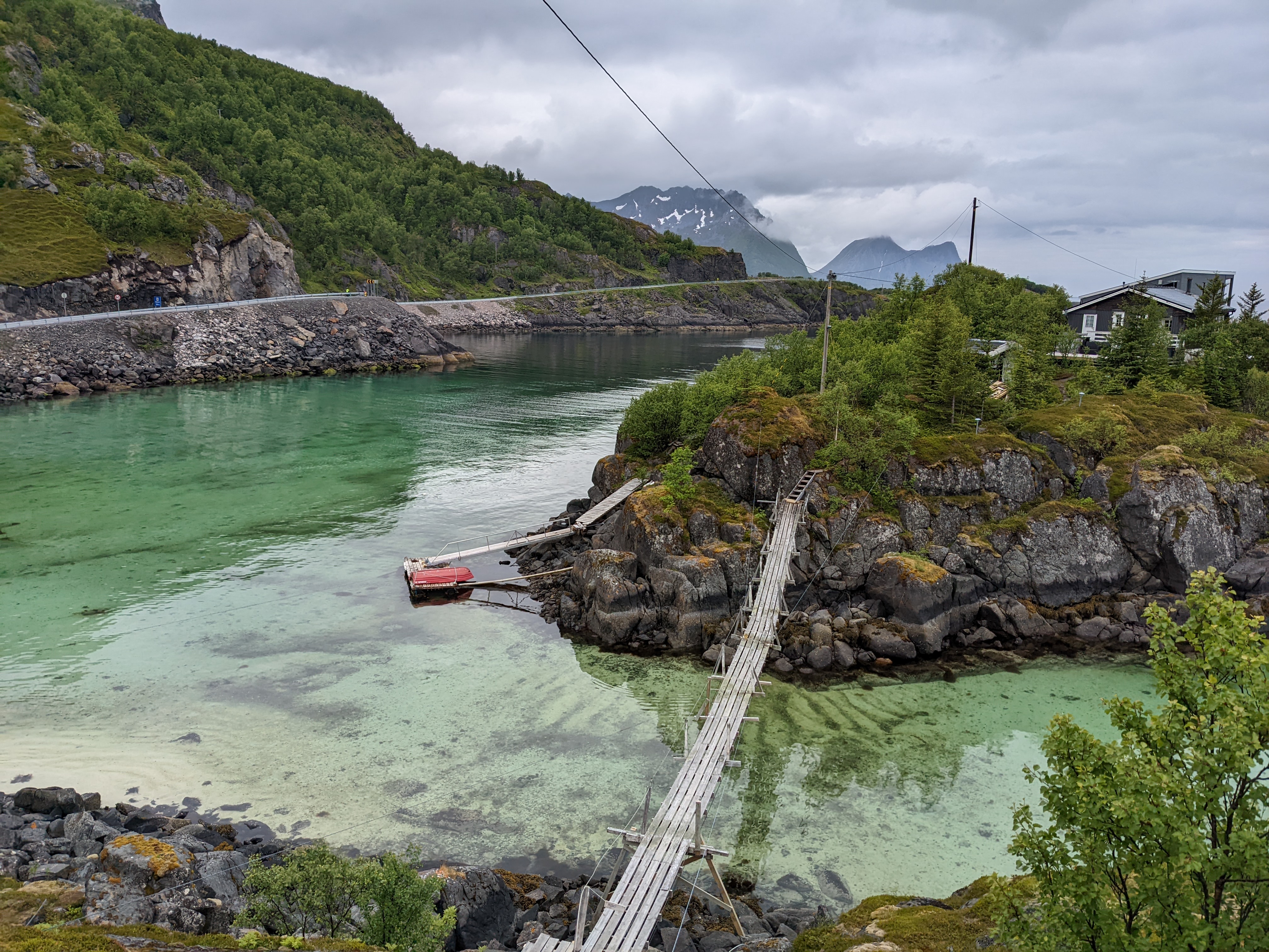 a narrow suspension bridge connecting a small island home with the mainland, over crystal-clear blue-green water above a white sand sea-floor