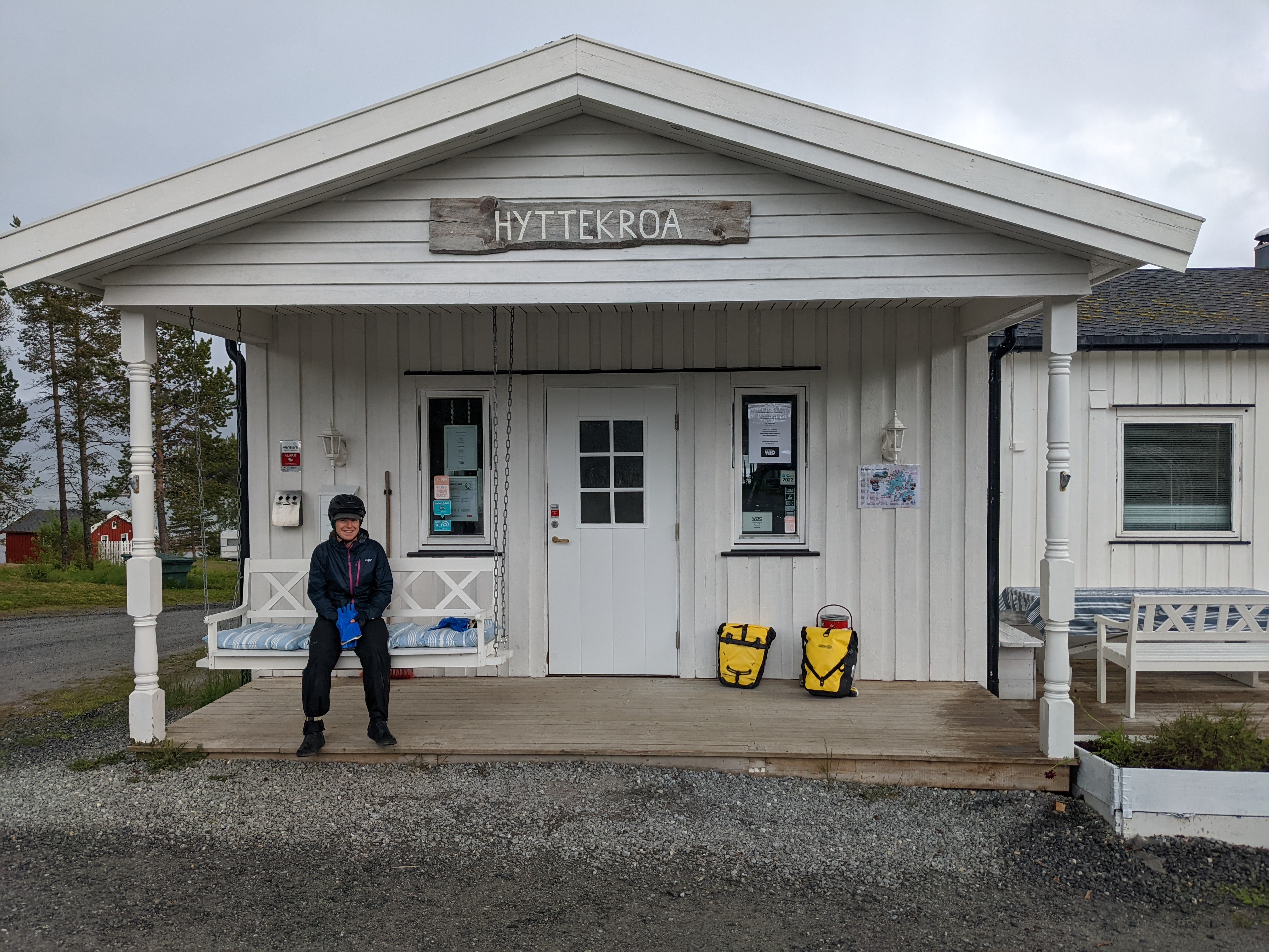 H sitting on a hanging bench in front of a white building wearing rain gear and a bike helmet