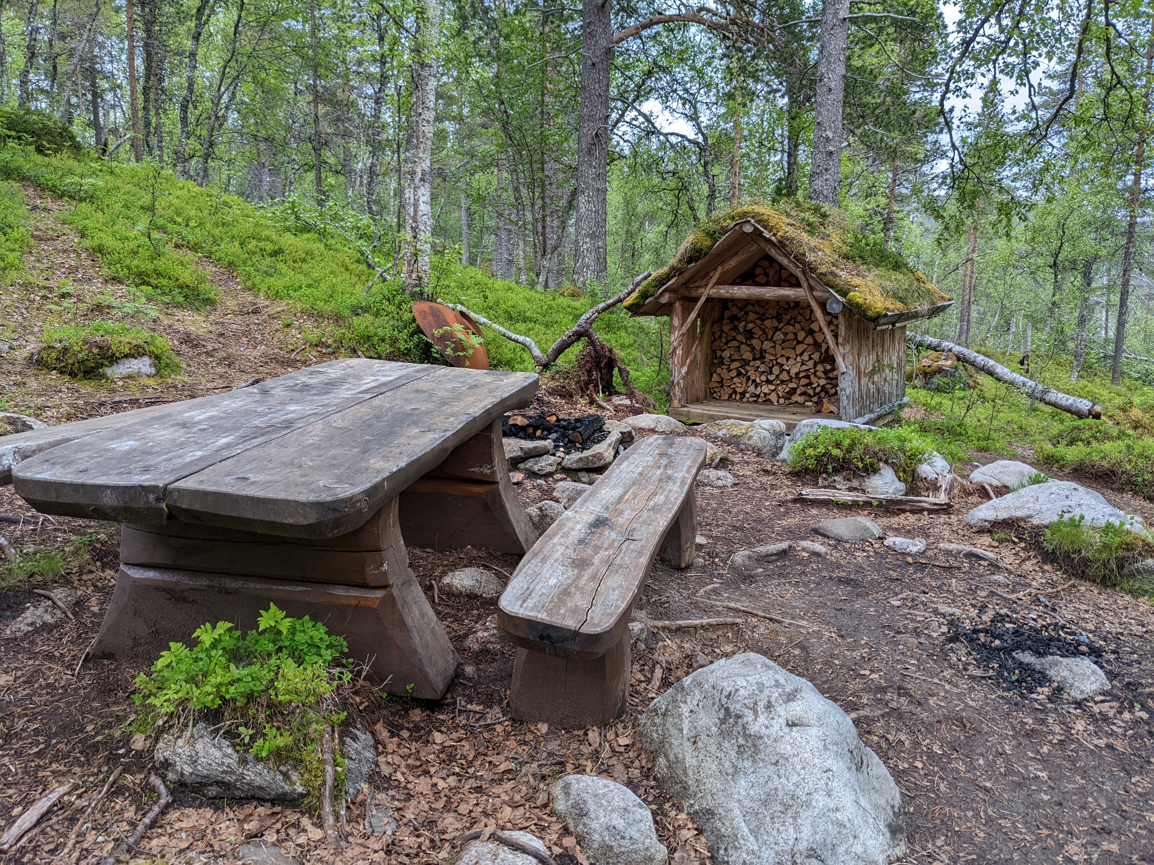 a heavy wooden picnic table in front of a grill, fire circle, and sod-rooved wood storage cabin