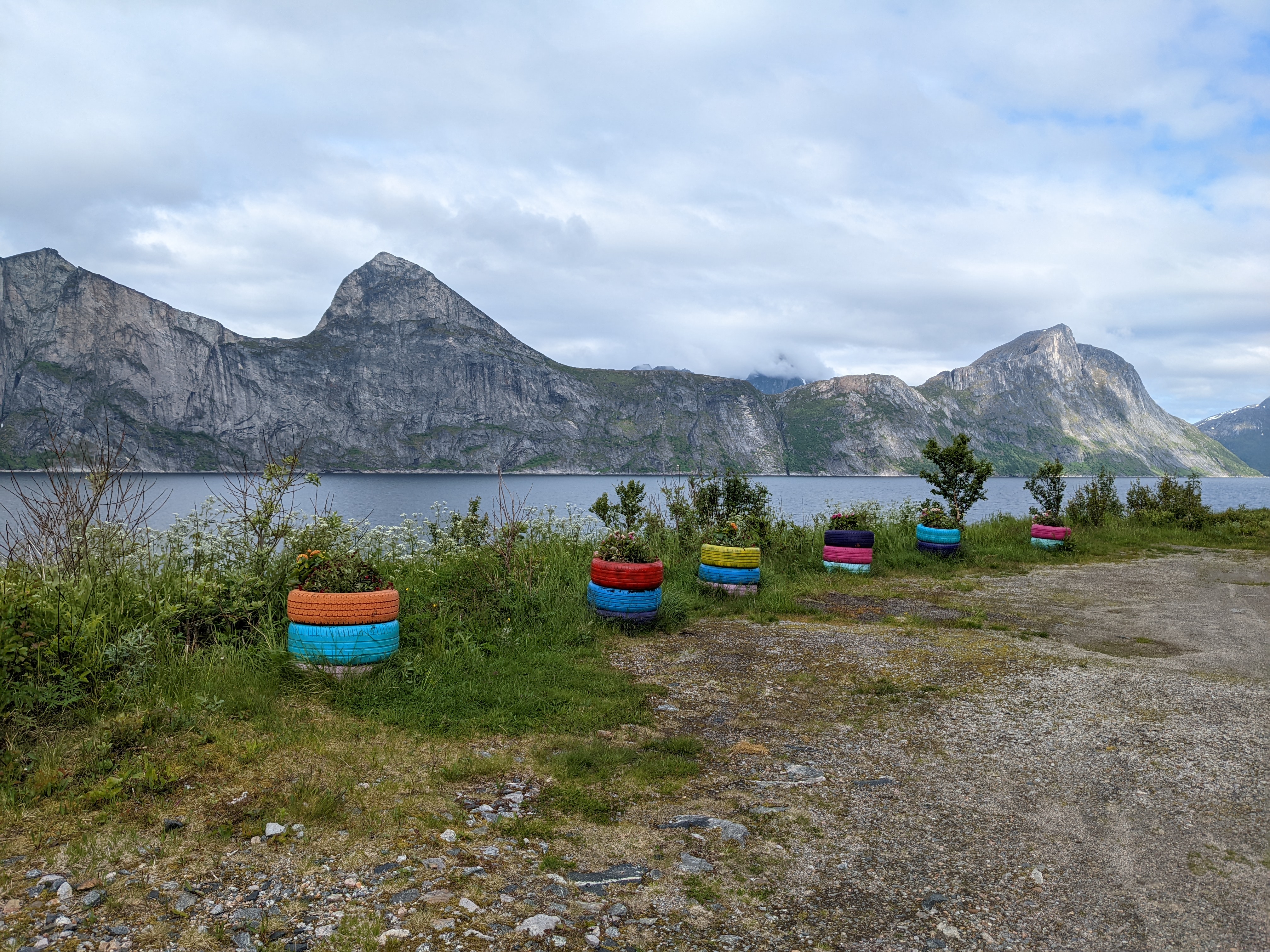 a row of sheer cliffs and peaks above the sea across a fjord, with funny colored-tire-stack planters in the foreground