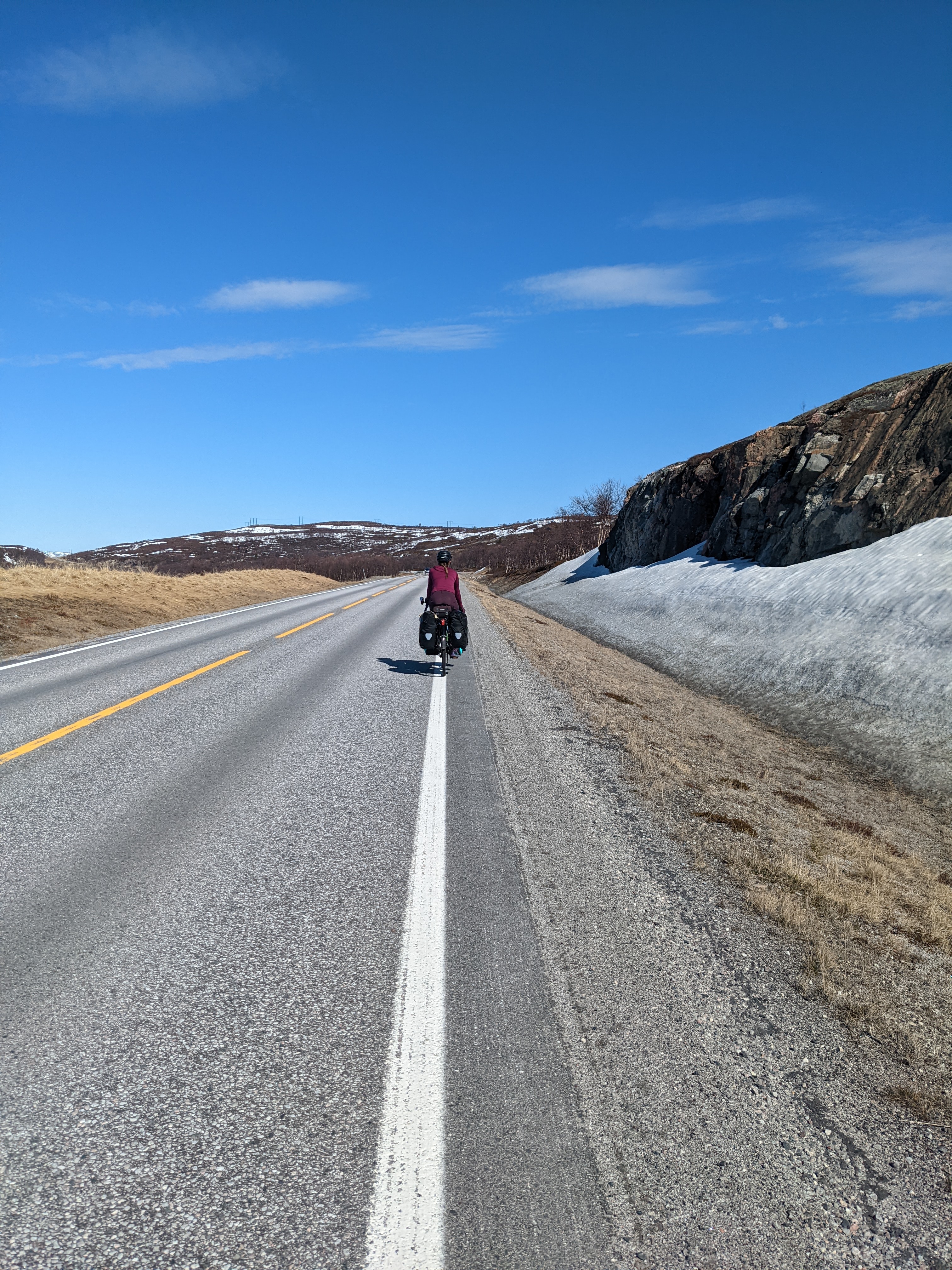 H riding down a straight road with brown plants on the roadside